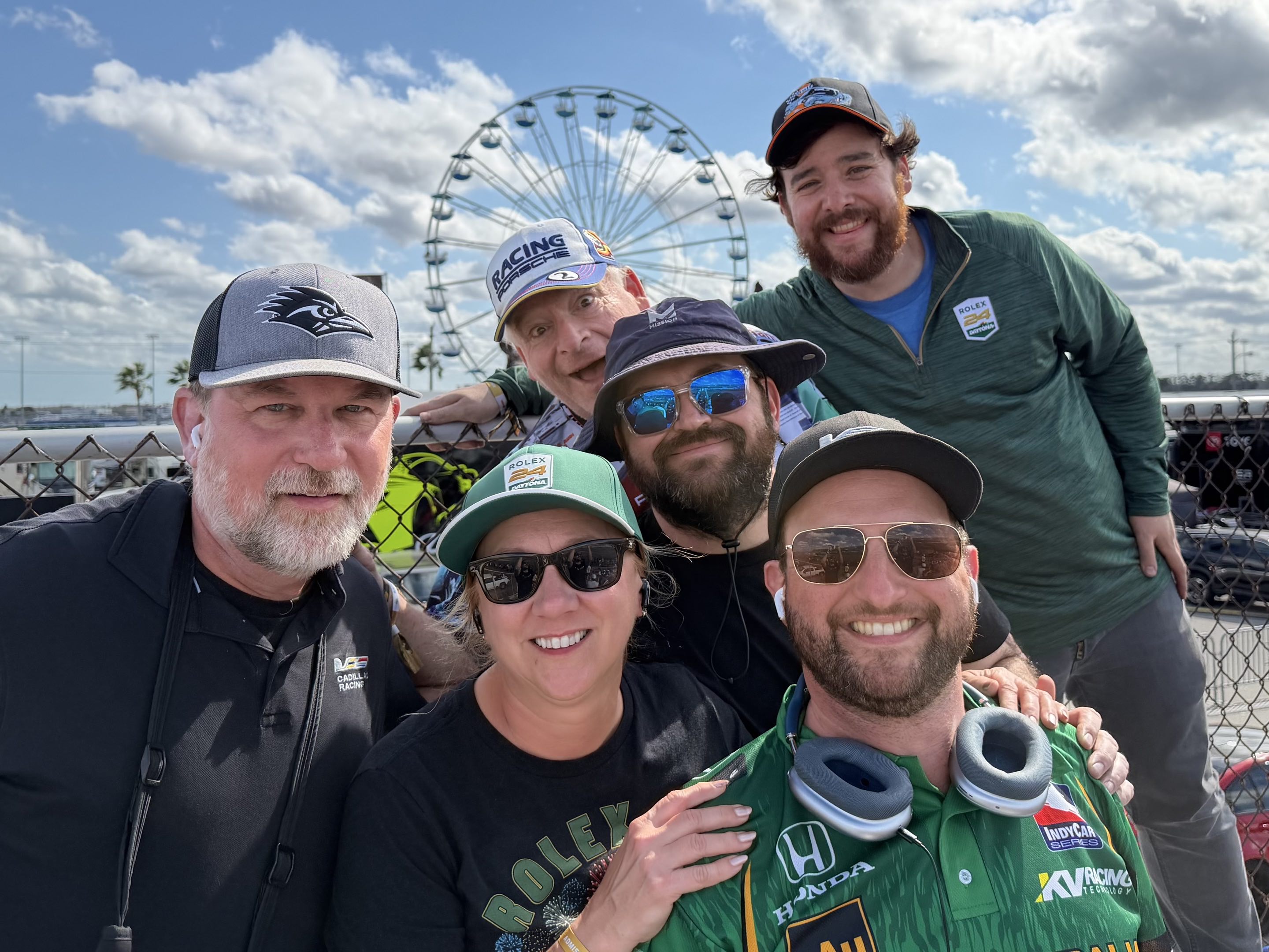 Six race fans smiling in front of the Daytona International Speedway ferris wheel in broad daylight after the Rolex 24 ended